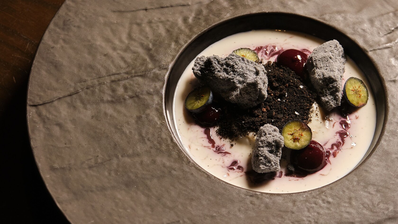 A bowl of panna cotta swirled with berry sauce, meringue pieces, chocolate crumble, whole cherries and sliced kumquats