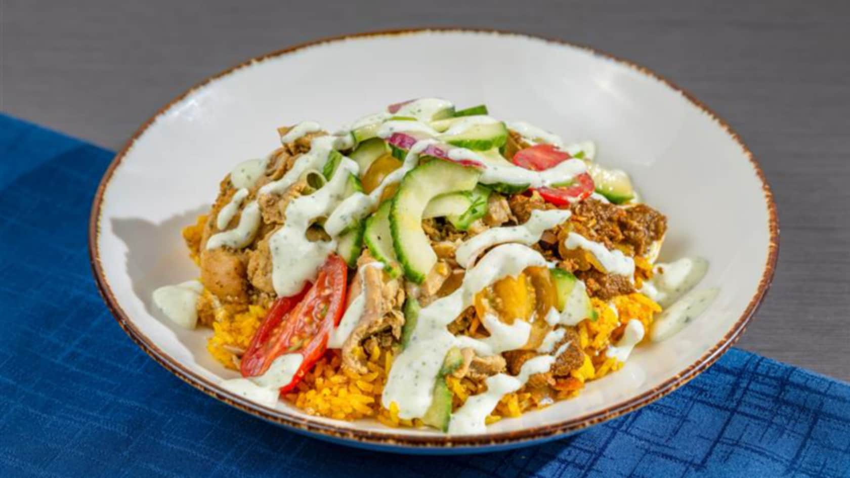 A beef and chicken bowl featuring rice, cucumber, tomato and tzatziki sauce