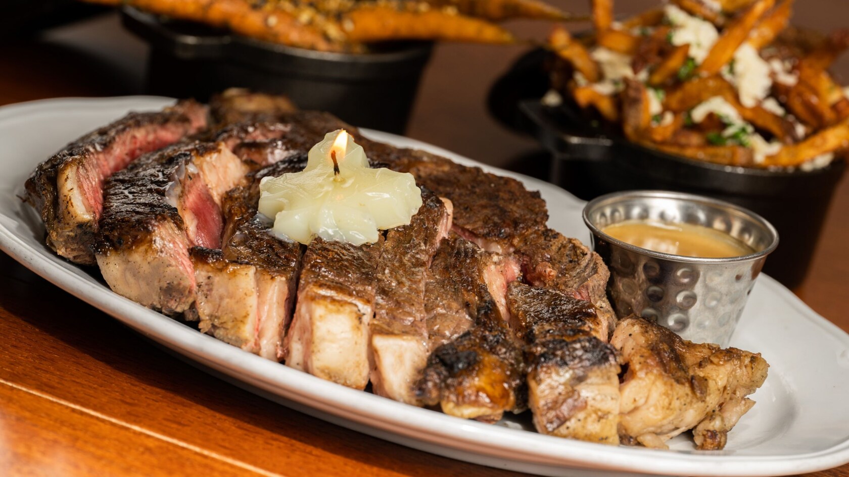 Un bistec grande con una vela de mantequilla y un ramequín de salsa, servido con acompañantes de poutine y zanahorias asadas