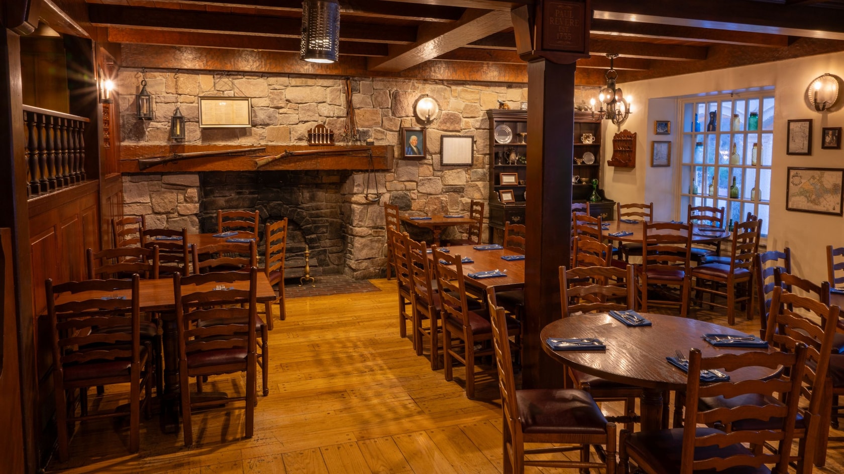 A rustic dining room with wooden chairs and tables, a stone fireplace with a wooden mantel and framed decor on the walls