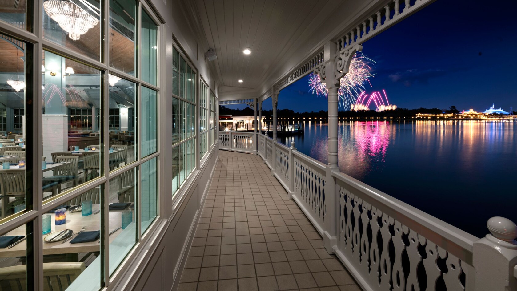 A restaurant balcony above a body of water, with lighted buildings and fireworks on the shore.
