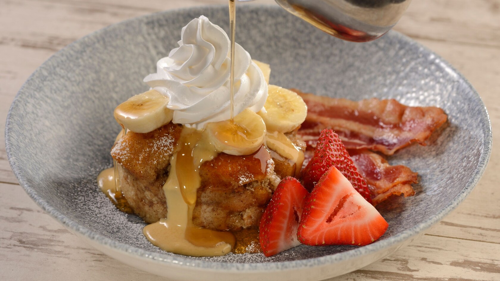 Syrup being poured over Banana Bread Pudding, strawberries and bacon