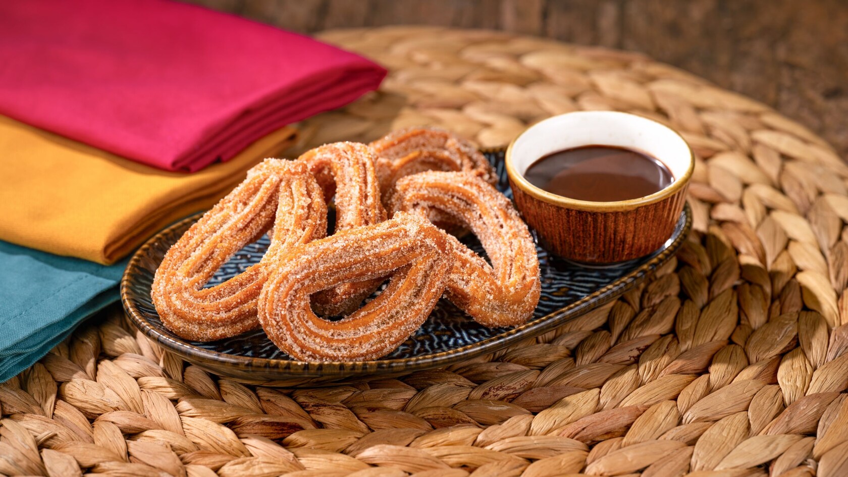 A plate of cinnamon sugar Churro Loops served with a cup of chocolate dipping sauce on a woven placemat