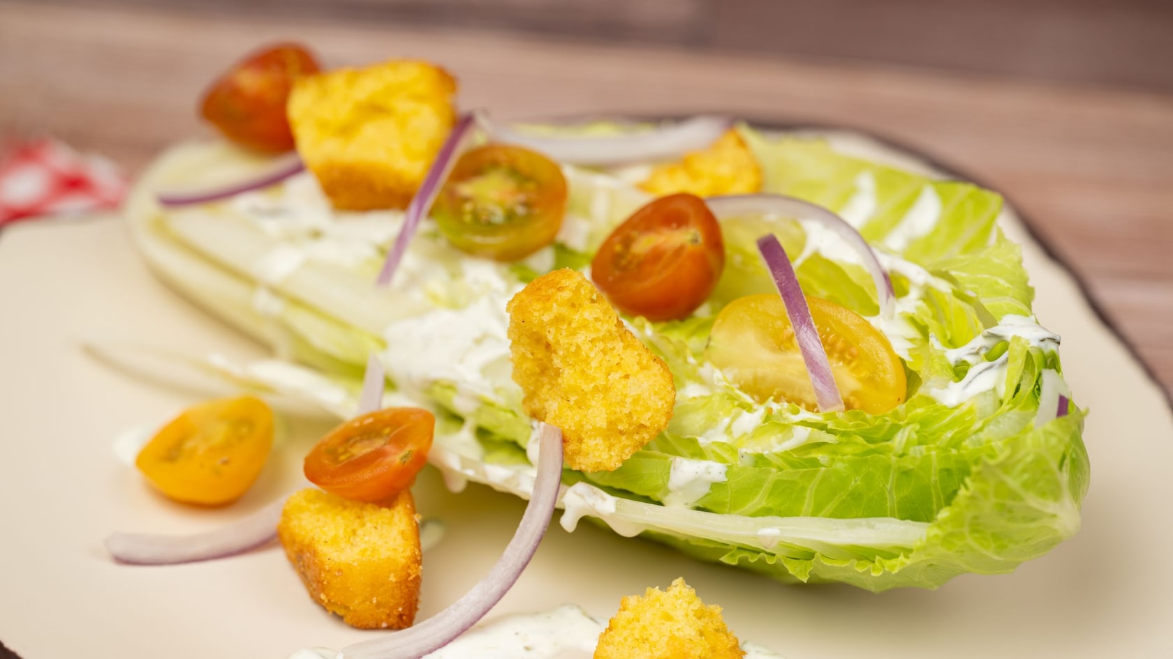 A salad featuring a wedge of romaine lettuce topped with creamy dressing, halved cherry tomatoes, thin slices of red onion and cornbread croutons