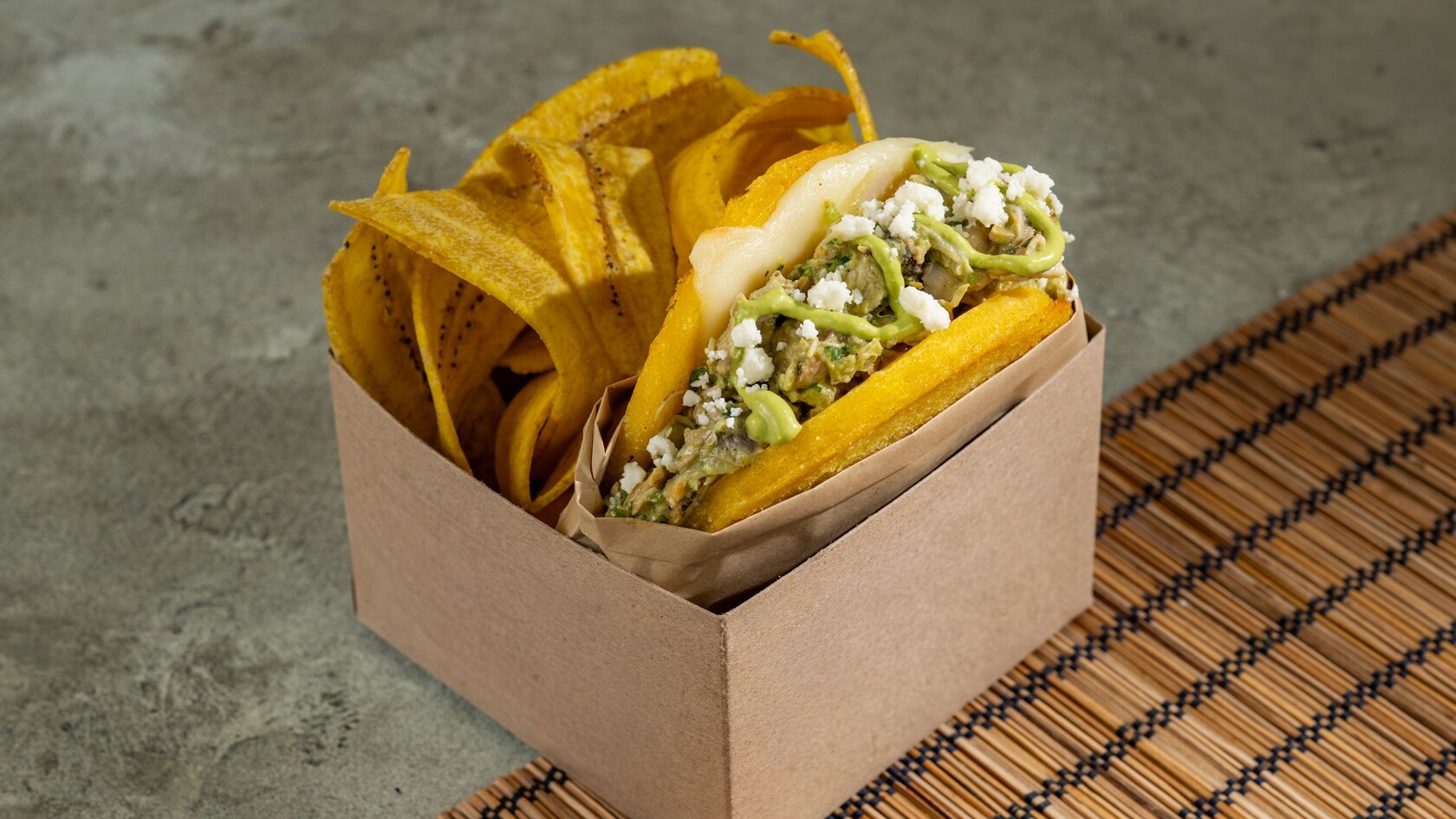 Shredded chicken in between 2 arepas and topped with sauce and cheese in a box with plantain chips