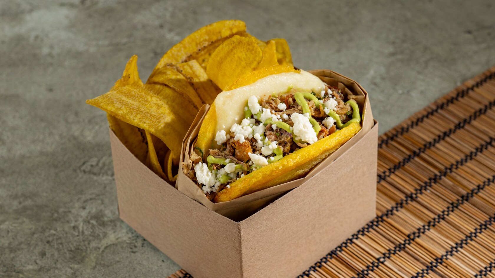 Shredded pork in between 2 arepas and topped with sauce and cheese in a box with plantain chips