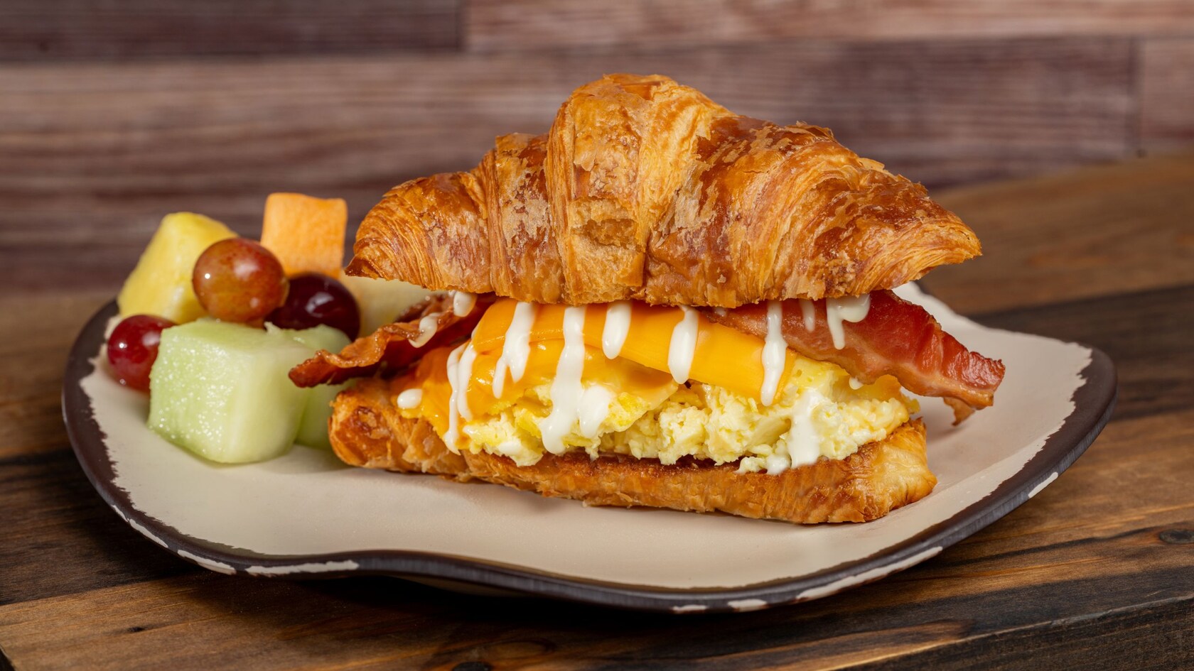 A croissant sandwich with eggs, cheese and bacon plated next to fruit salad from Sunshine Seasons