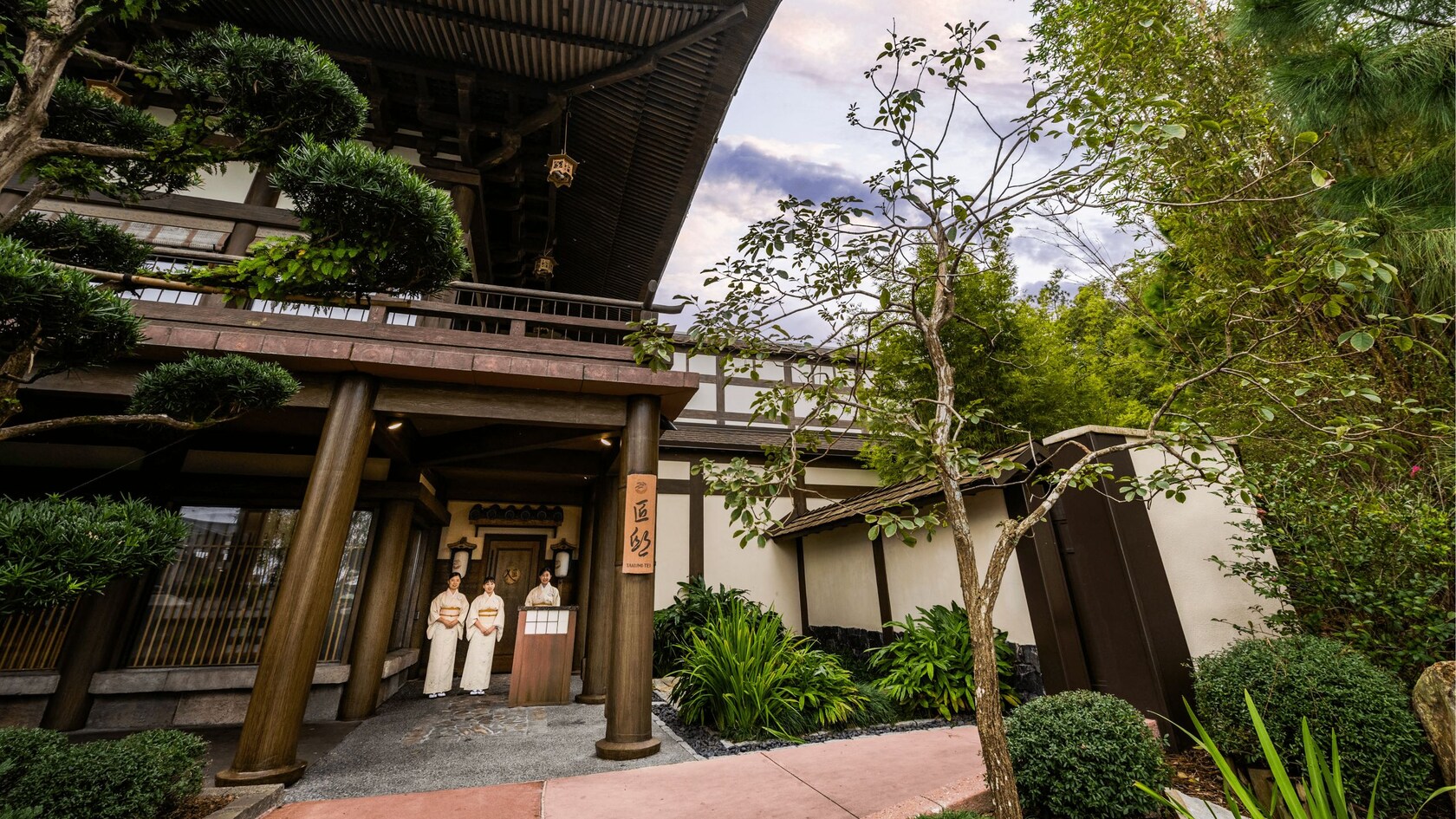 Three Cast Members dressed in kimonos standing under the entrance of a pagoda at Takumi Tei
