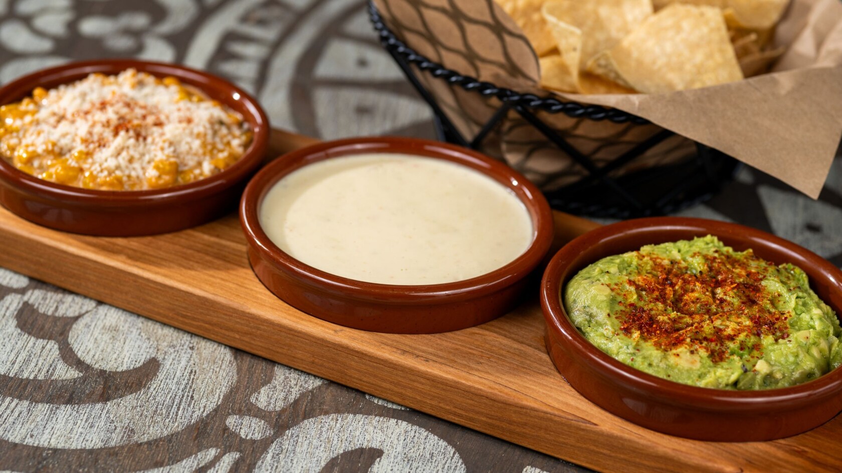 A basket of chips next to a serving board with roasted corn, queso and guacamole dips