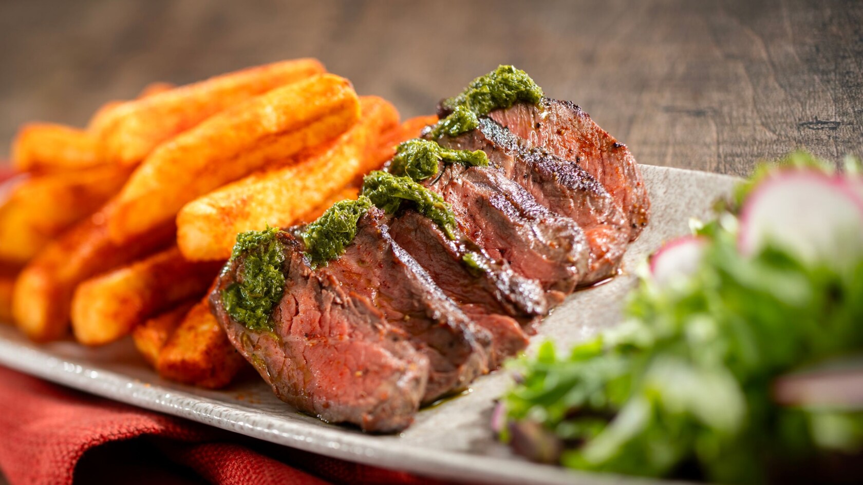 Bistec en rodajas cubierto con salsa chimichurri, servido con papas fritas sazonadas y acompañante de ensalada en un plato 