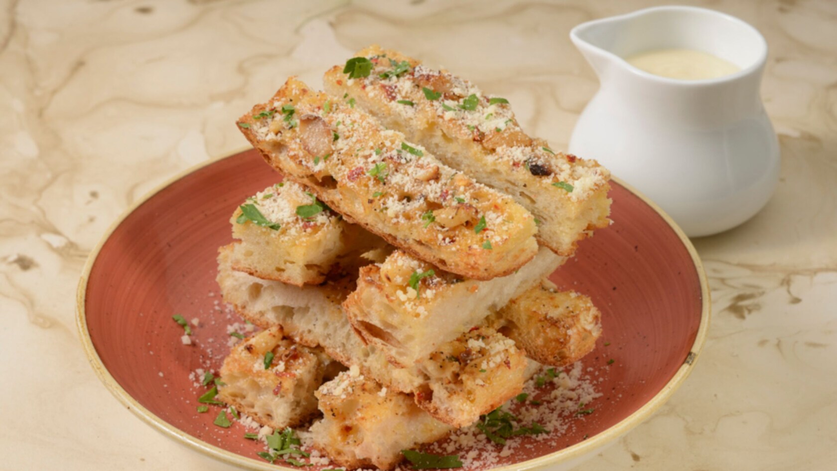 Slices of garlic bread are stacked on a dish, next to a small vessel of parmesan fonduta
