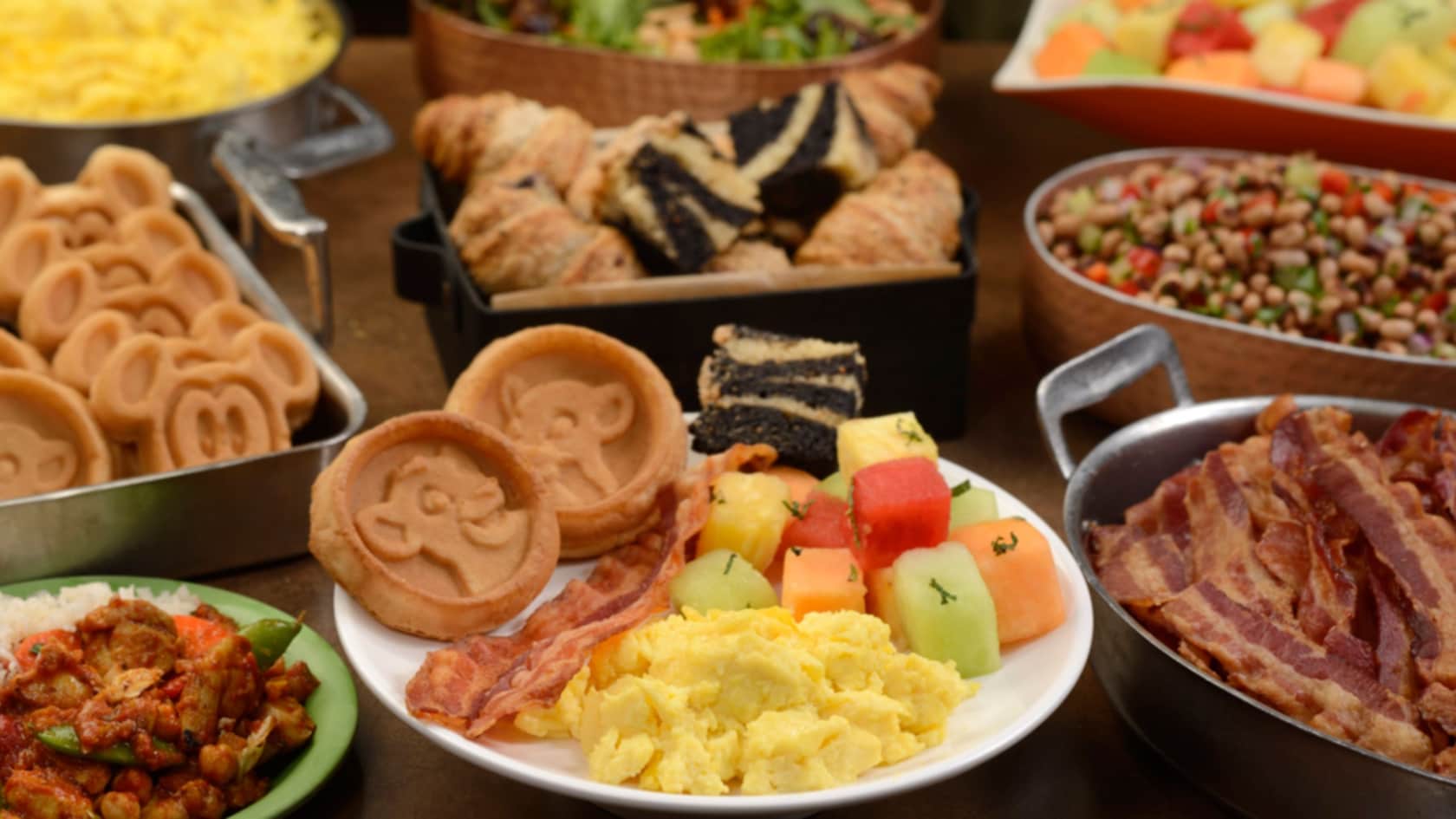 Several platters of breakfast foods, including Mickey waffles, bacon, eggs, pastries and fresh fruit