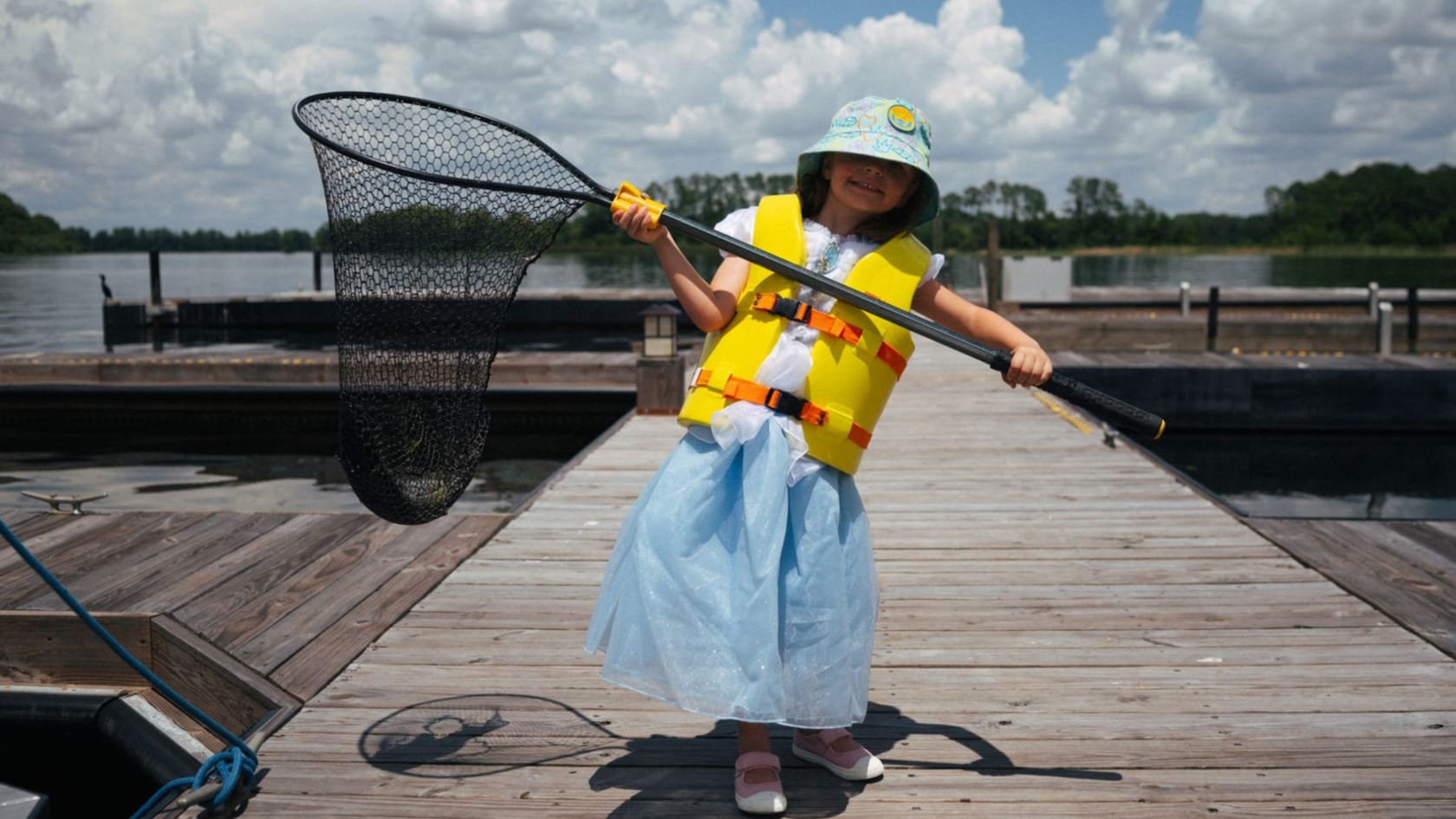 A young girl with a fishing hat and a life vest stands on a dock with a fishing net that holds her catch