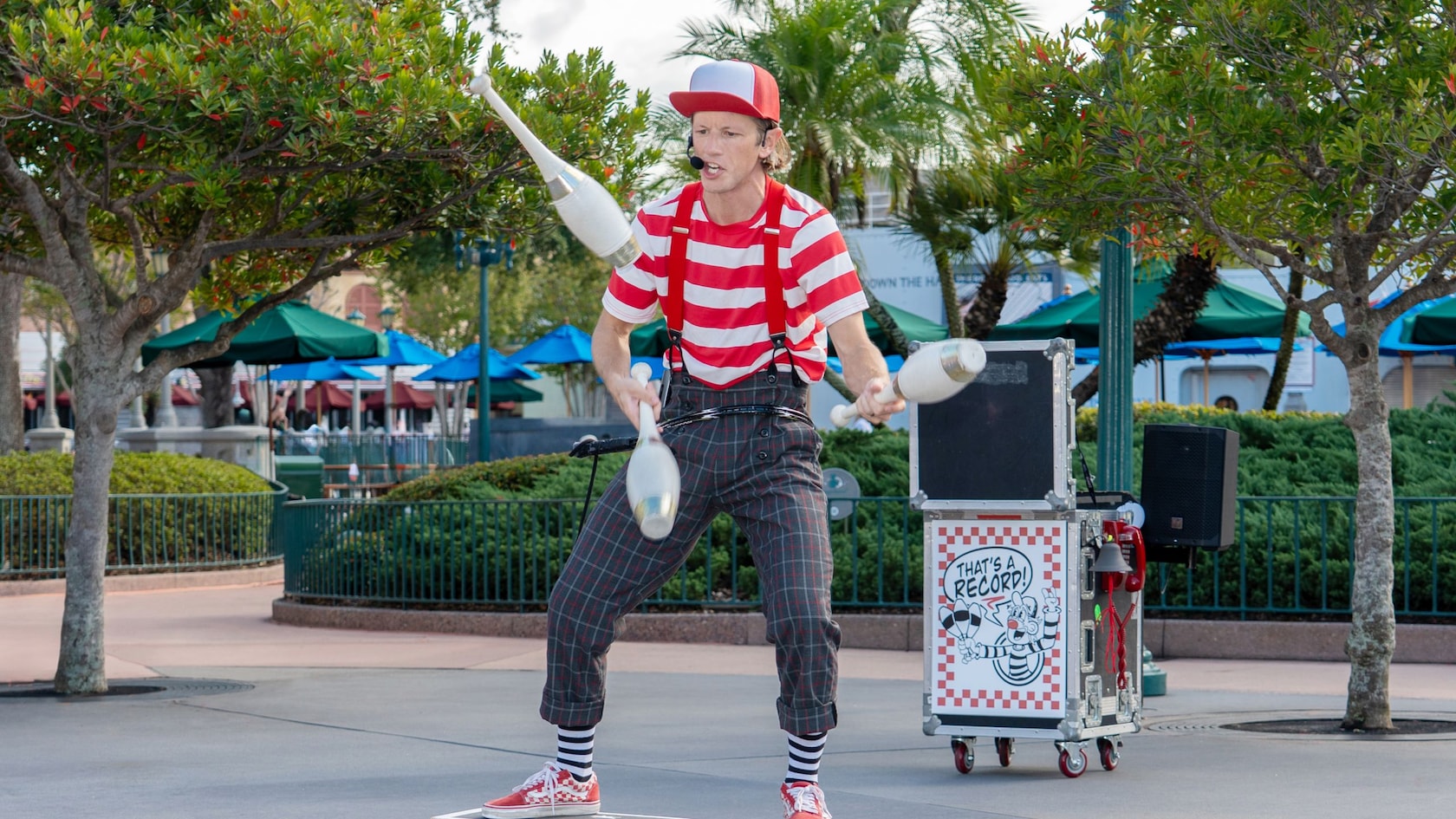 A man in a striped shirt, plaid pants and wide suspenders juggles 3 clubs and teeters on a balance board