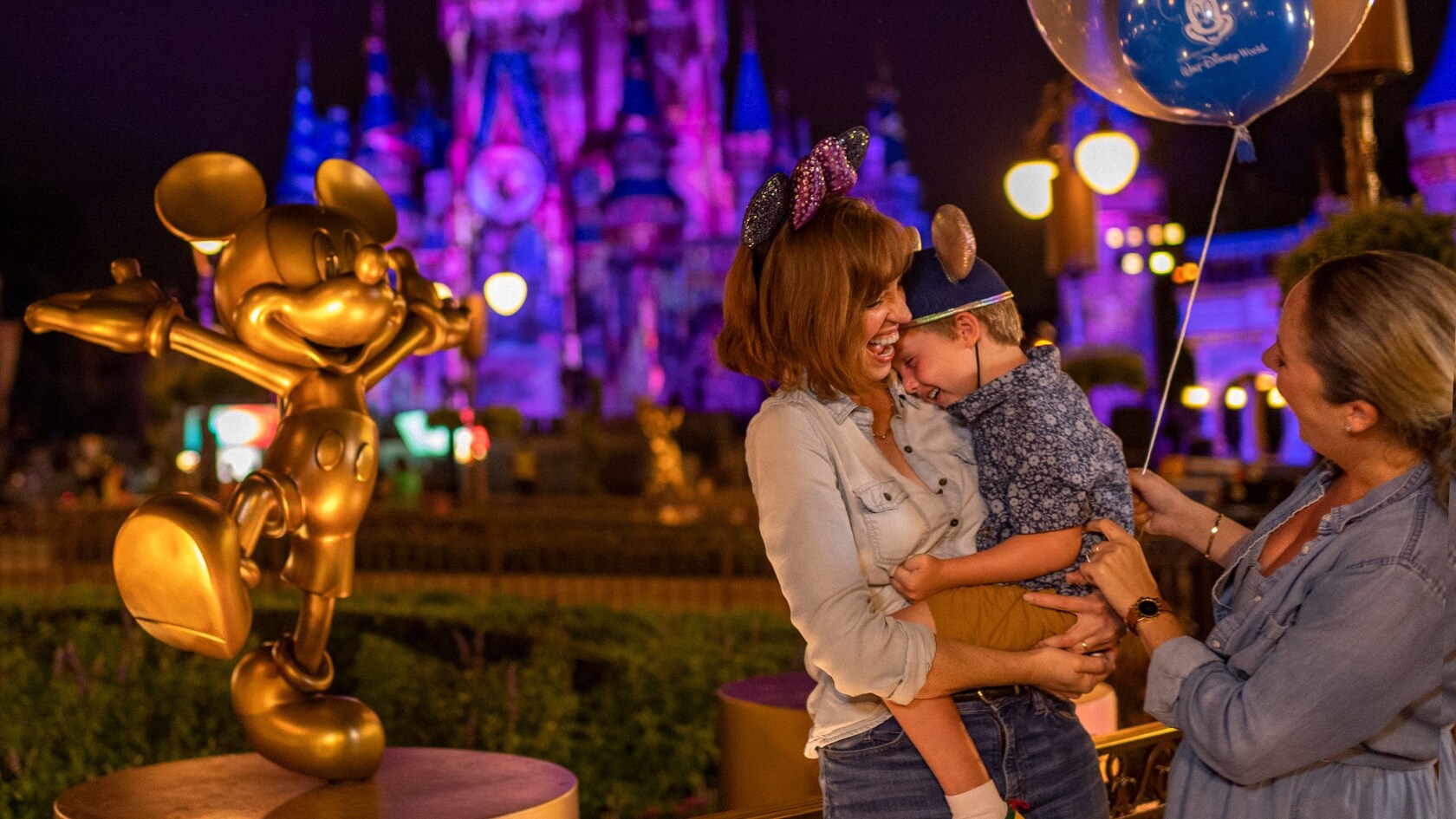 A mother holds her son and laughs while another woman holds a balloon near a Mickey Mouse statue near Cinderella Castle 