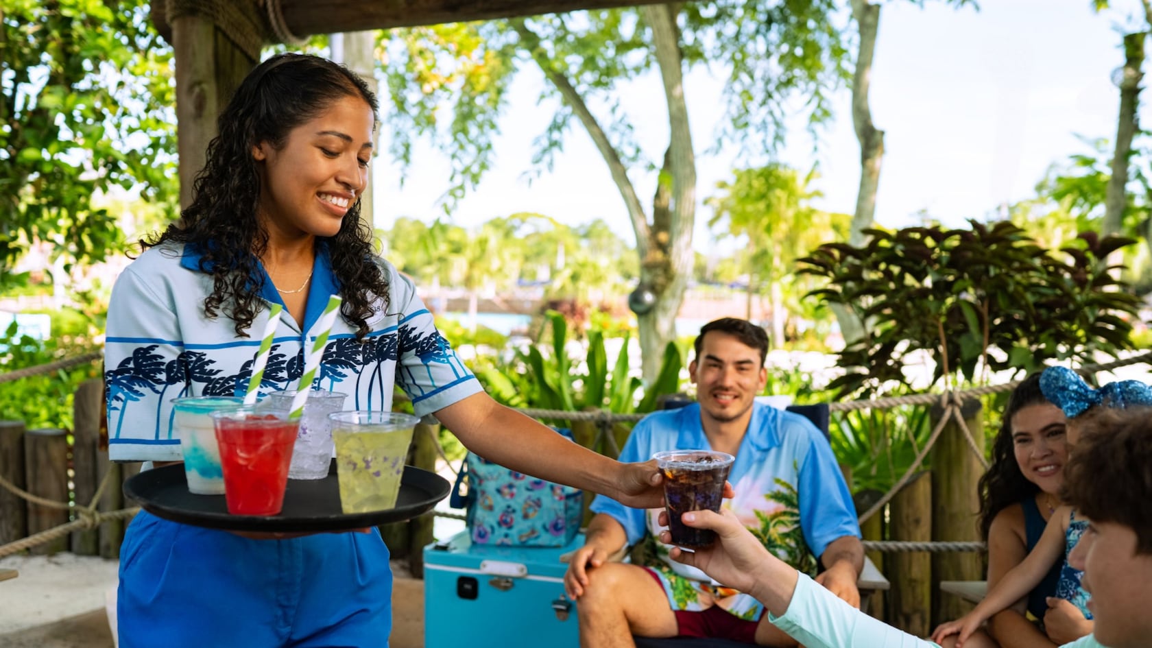 A Cast Member serving tropical drinks to a family relaxing in a shaded beach cabana at Disney’s Typhoon Lagoon water park