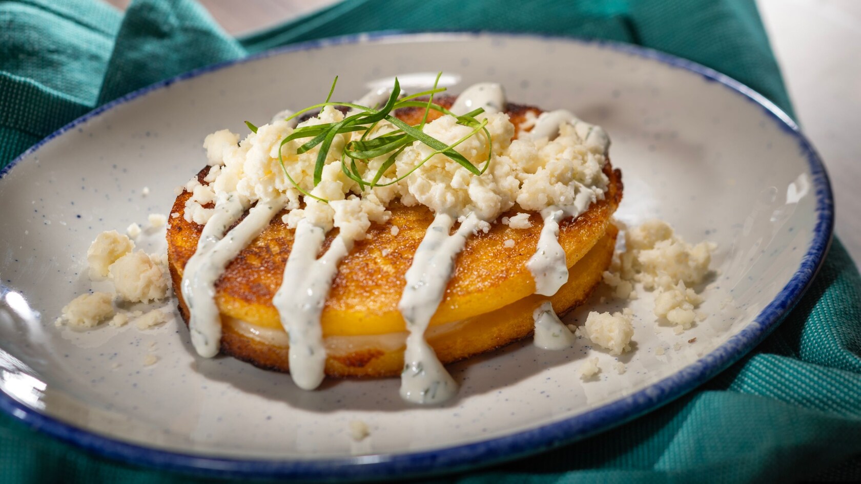 Una arepa rellena de queso, cubierta con salsa blanca, queso desmenuzado y hierbas