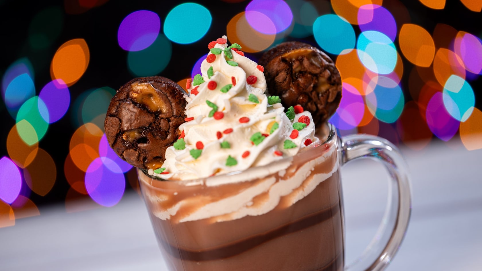 A glass of hot chocolate topped with whipped cream, sprinkles and 2 chocolate cookies