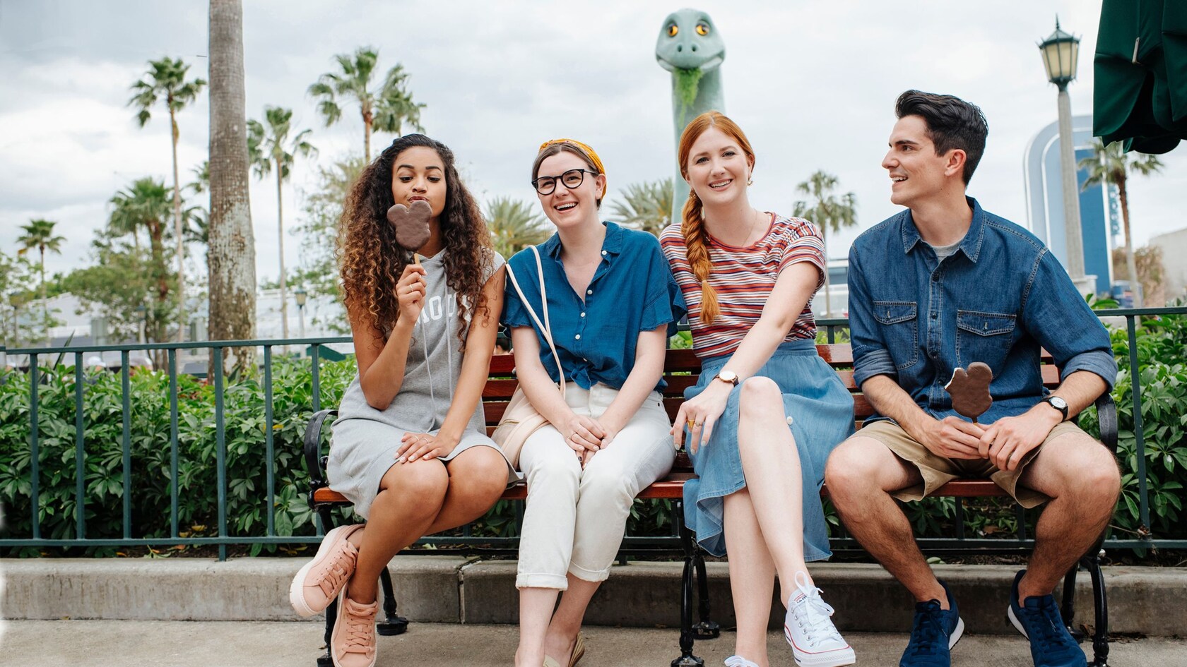 Quatre amis, deux avec des barres de crème glacée Mickey, sont assis sur un banc devant Gertie le dinosaure à Disney’s Hollywood Studios