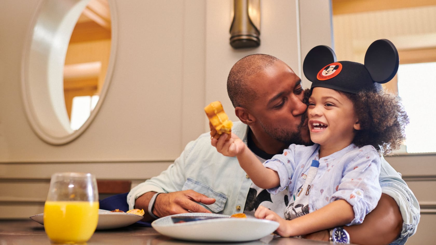 A man kisses a child on a cheek while sitting at a table with orange juice and Mickey waffles