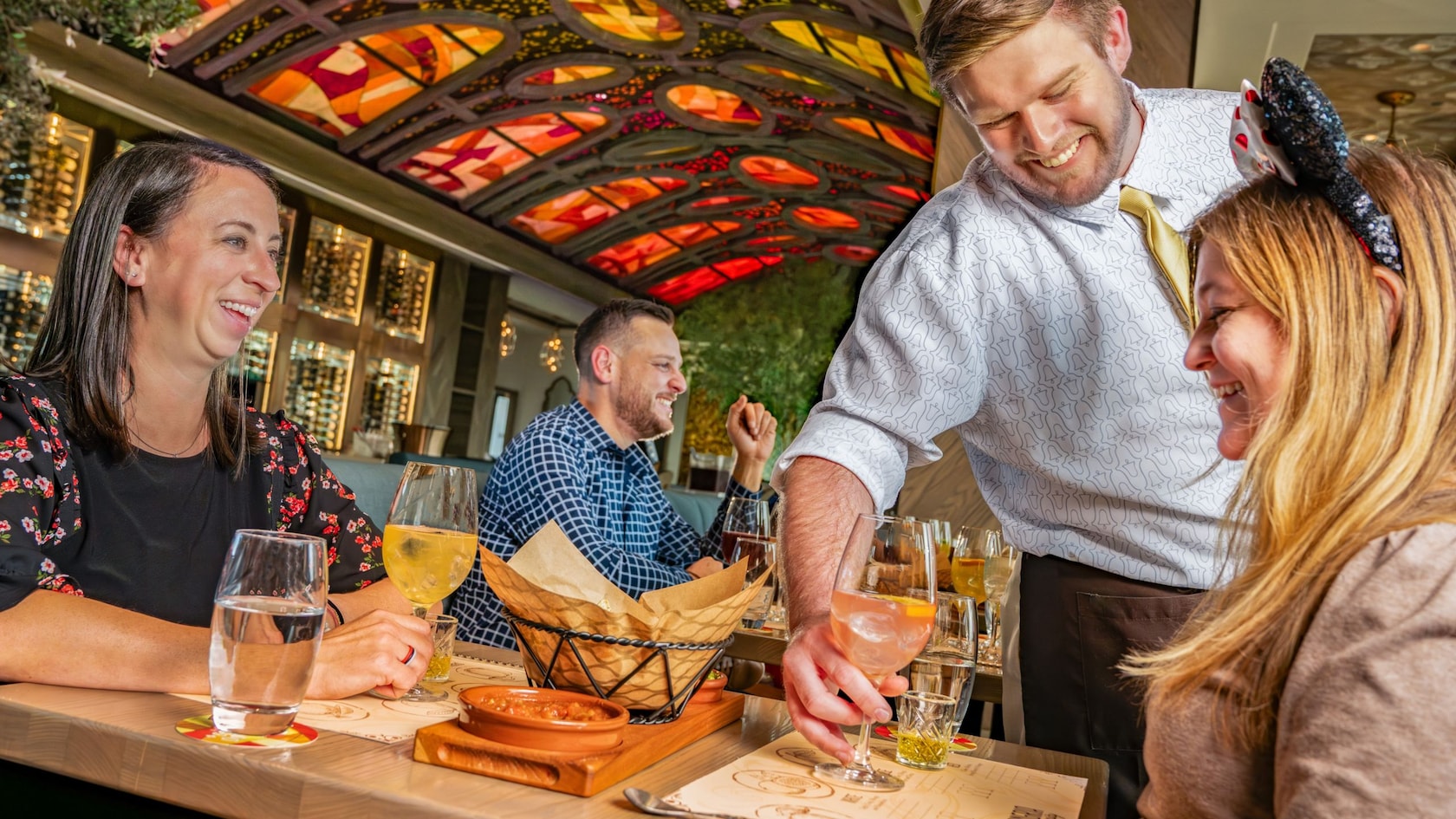 A Disney Cast Member serving a beverage to a woman who is sitting with a friend at a tasting class