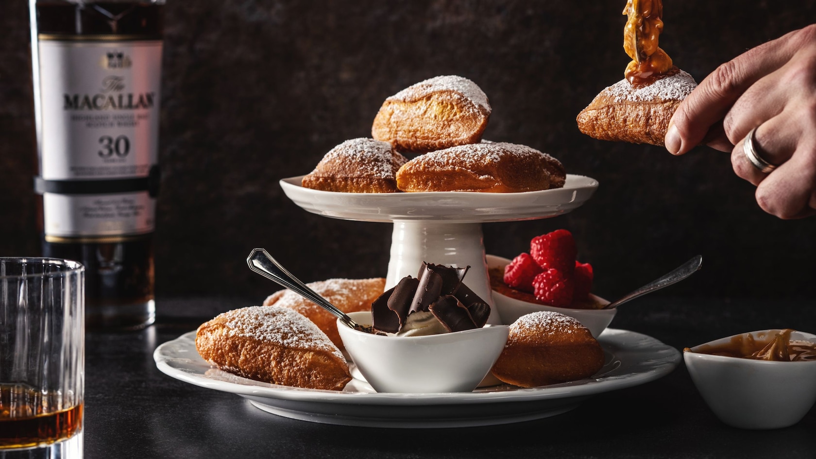 Un plato de varios niveles con beignets y pequeños tazones con ingredientes junto a una botella y un vaso de whisky escocés The Macallan