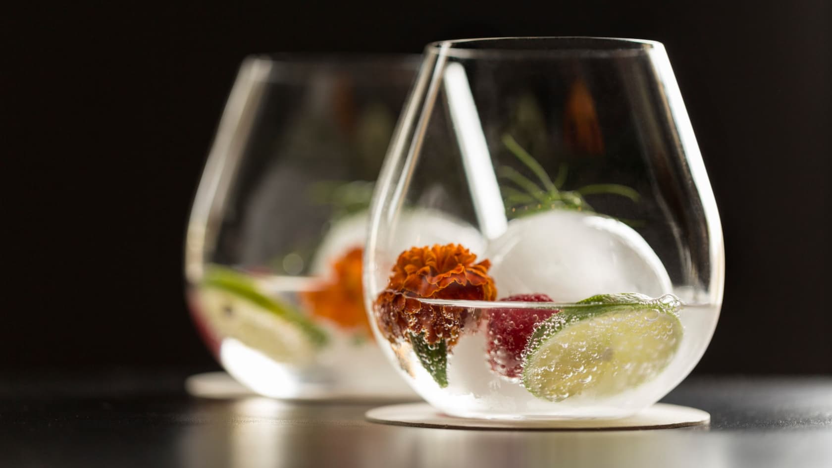 Two stemless glasses filled with cocktails and garnished with lime, berries, rosemary and ice