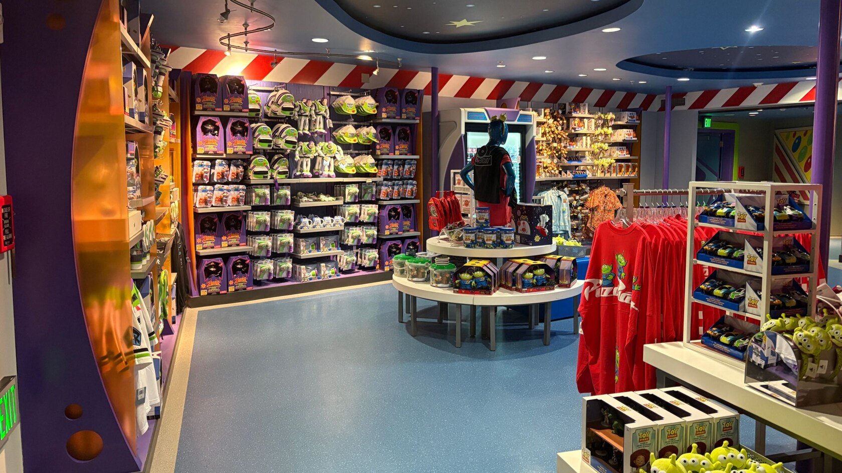 Interior of the brightly lit Buzz Lightyear Shop in Tomorrowland with blue floors and curved ceiling panels, featuring shelves and tables filled with colorful merchandise—plush toys, boxed items, and clothing—from Toy Story.