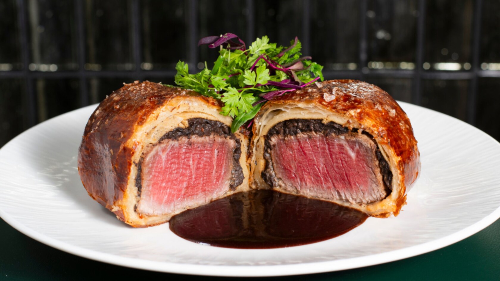Beef Wellington sliced open to reveal a beef fillet encased in a mushroom duxelles and puff pastry, served on a plate with a jus and a garnish of microgreens