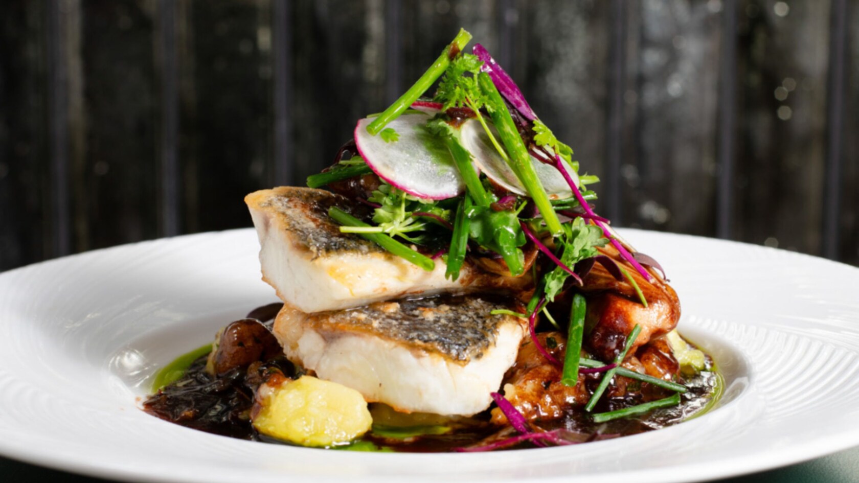 A branzino fillet stacked over mushrooms and a sauce, topped with radish slices, chives, red cabbage microgreens and herbs