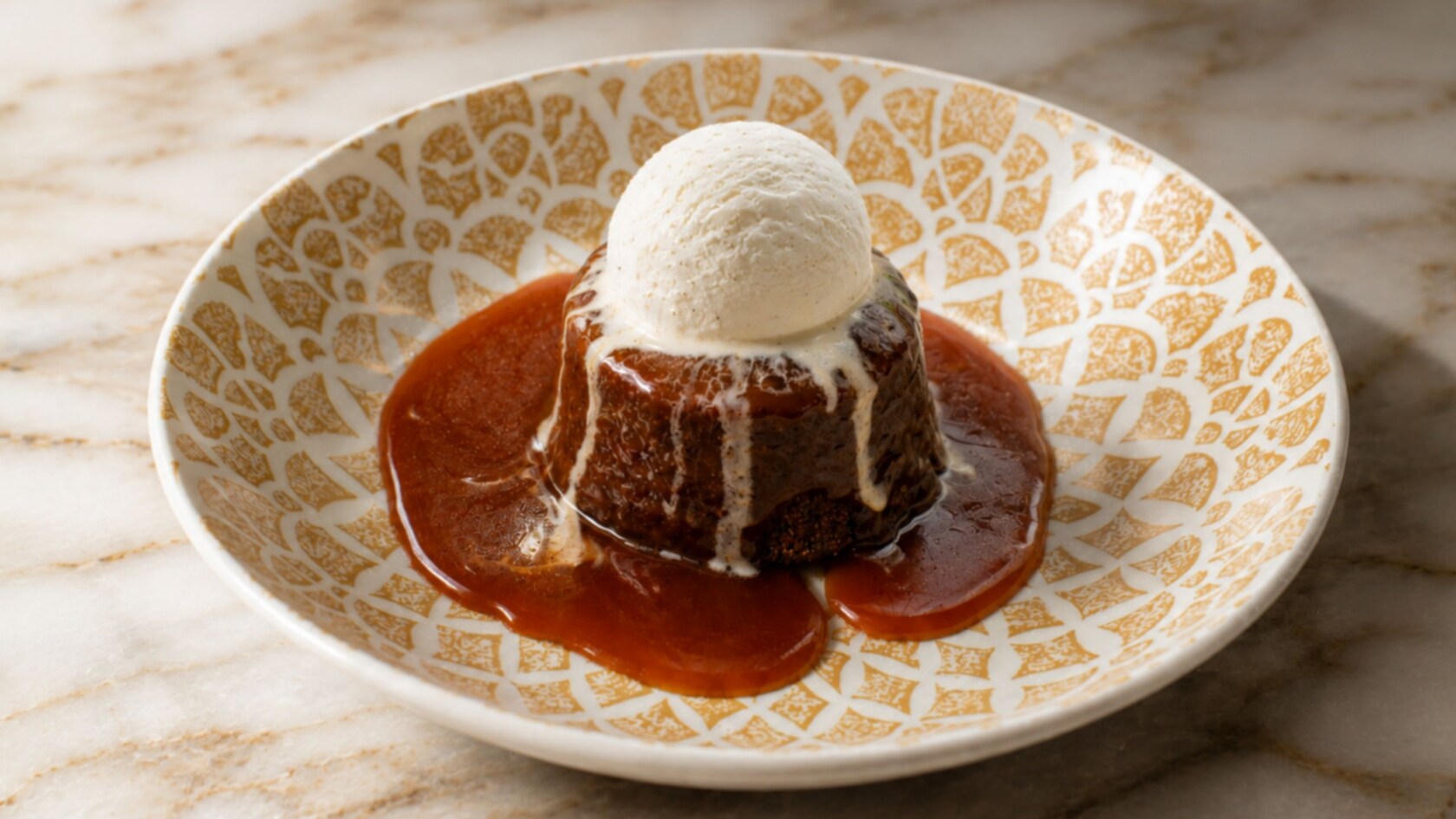 A sticky toffee pudding served in a bowl with toffee sauce and topped with a scoop of vanilla ice cream
