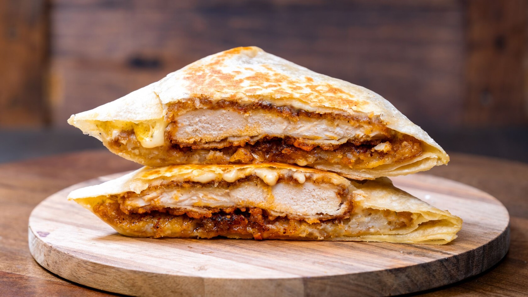 Both halves of a folded chicken sandwich with melted cheese served on a round wooden board