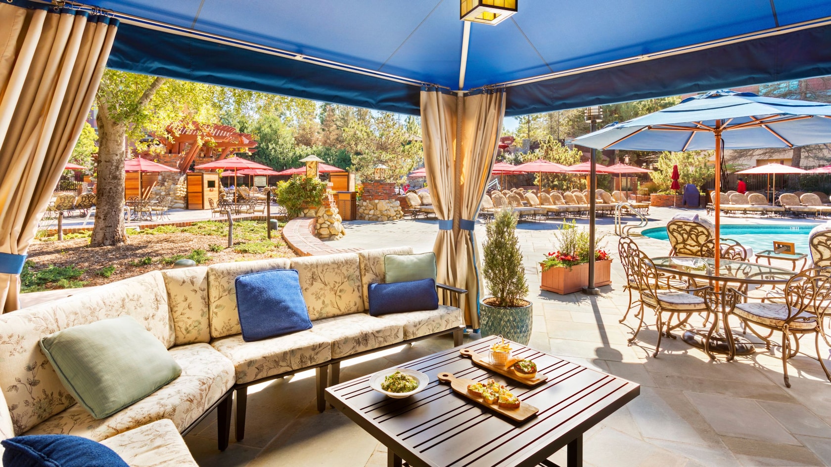 A large poolside cabana with a sofa, ceiling fan and a table with snacks