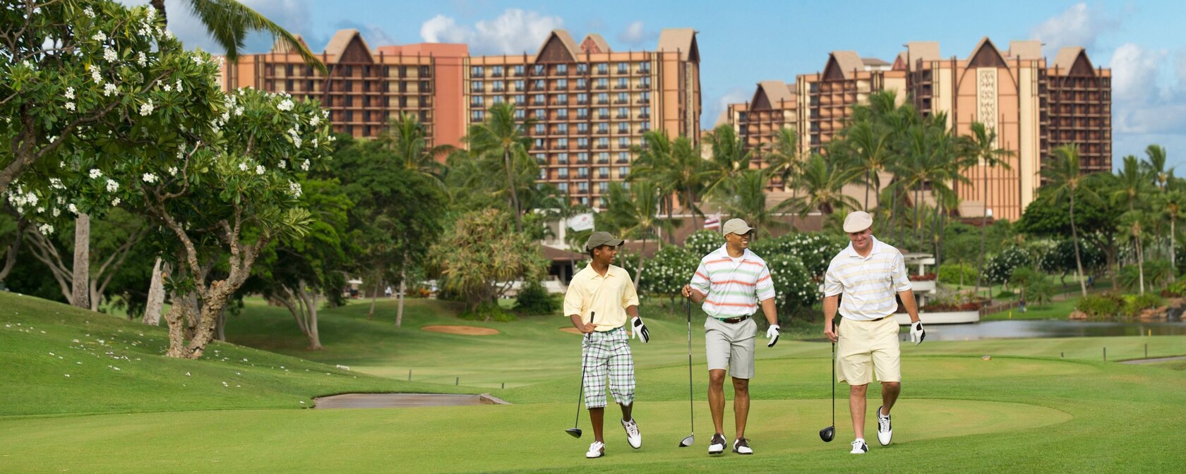 3 men wearing shorts and caps walk down a lush golf course in front of a large hotel