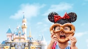 Una niña parada frente al castillo Sleeping Beauty Castle porta un sombrero con orejas de Mickey Mouse y sostiene un pretzel