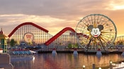 A bridge across a waterway leads through an arch that marks the entrance to Paradise Pier, an area featuring a carousel, a large wooden rollercoaster and a Ferris wheel with Mickey Mouse's face at the center