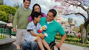 A child with a plush Donald Duck toy sits on the lap of a man in a wheelchair with his parents smiling behind in front of the Magic Castle