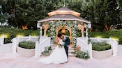 A couple in wedding attire stand in front of a gazebo adorned with flowers and decorative lanterns