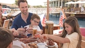 A young girl cracks a lobster claw, as her brother and father sit with her at a dockside table containing a beer and a plate of lobster