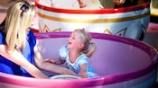 A laughing mother and toddler daughter sit in a spinning oversized teacup