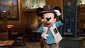 Mickey Mouse, dressed as an explorer, standing in a large dining room furnished in the style of a wilderness lodge
