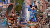 Mickey Mouse in wizard attire stands atop a float in a parade marching near Sleeping Beauty Castle