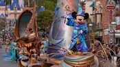 Mickey Mouse in wizard attire stands atop a float in a parade marching near Sleeping Beauty Castle