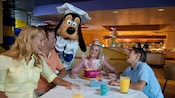 A family smiles at a table while Goofy laughs nearby in a chef costume