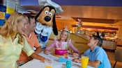 高飞（Goofy）在一个餐厅里在一群微笑的家人旁摆姿势