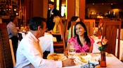 A man and woman smile at a table plated with food in a bustling restaurant