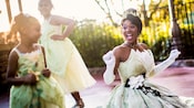 Princess Tiana in a fenced courtyard laughs with 2 girls wearing Princess Tiana costumes
