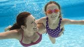 A mother and daughter wearing goggles swim underwater in a swimming pool
