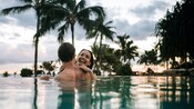 海岸線沿いに並ぶヤシの木のシルエットを背景に、インフィニティ・プールで笑い合う 2 人の人たち 海岸線沿いに並ぶヤシの木のシルエットを背景に、インフィニティ・プールで笑い合う 2 人の人たち