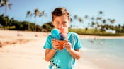 A boy stands on a tropical beach near the ocean and holds a bucket containing a Mickey Mouse balloon